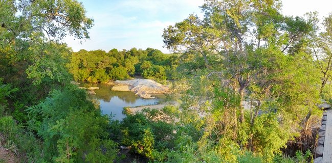 Panoramic view of a tranquil waterhole in Kruger National Park showing large granite rock formations rising from calm water, surrounded by dense green acacia trees and bush vegetation under golden afternoon sunlight