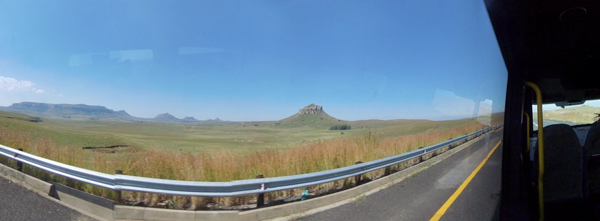 Panoramic landscape photograph taken from inside a vehicle showing a paved road with metal guardrail, golden-brown grassland plains, and rocky mountain formations in the distance under a clear blue sky