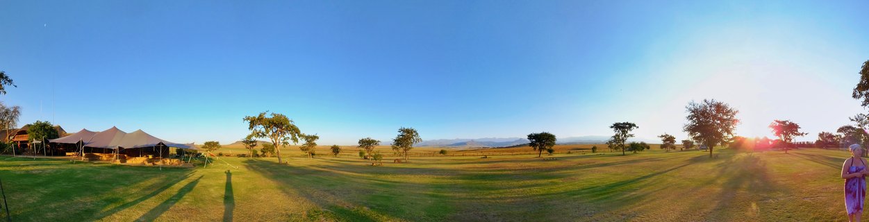 Wide landscape showing safari lodge tents on grassy field with scattered trees and mountains in background under clear blue sky at sunset in South Africa