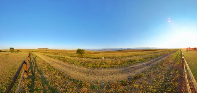 Panoramic view of a Y-shaped dirt road junction through golden grassland fields with wooden fences on both sides, scattered trees, and distant mountains under a clear blue sky at sunset