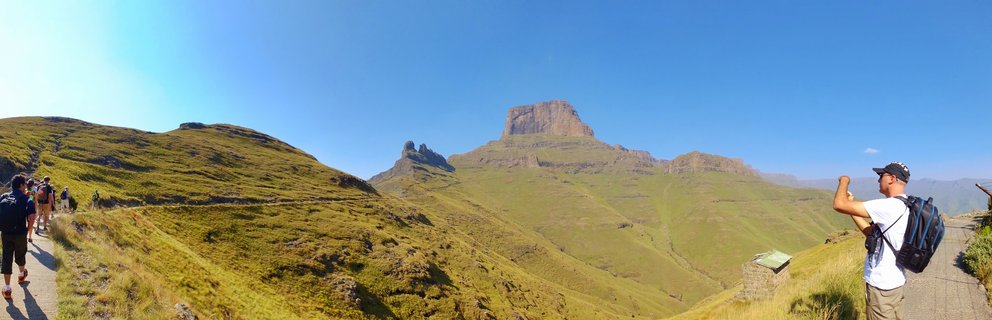 Panoramic view of hikers on a mountain trail with green slopes and flat-topped Drakensberg peaks under blue sky in South Africa