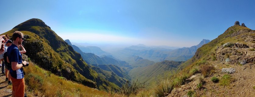 Group of hikers at mountain viewpoint overlooking expansive Drakensberg valley with layered mountain ranges, green slopes, rocky outcrops, and clear blue sky in South Africa