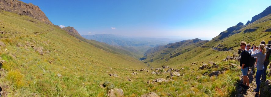 Panoramic view of green mountain valley in Drakensberg with rocky cliffs on both sides and a group of hikers standing on a trail on the right, distant mountain ranges visible under blue sky