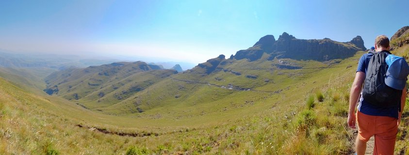 A hiker with a black backpack and orange shorts stands on a grassy mountainside looking out over a green valley toward rocky mountain peaks under a clear blue sky in Northern Drakensberg, South Africa