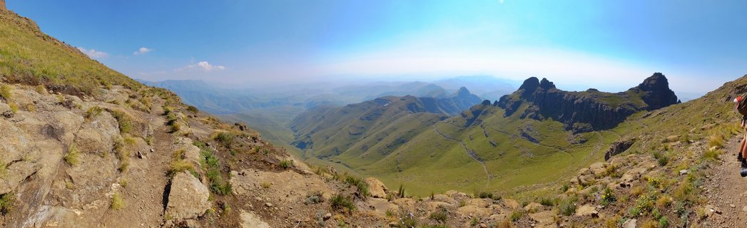 Wide panoramic mountain landscape showing green Drakensberg valley with rocky peaks, hiking trail, and blue sky in South Africa