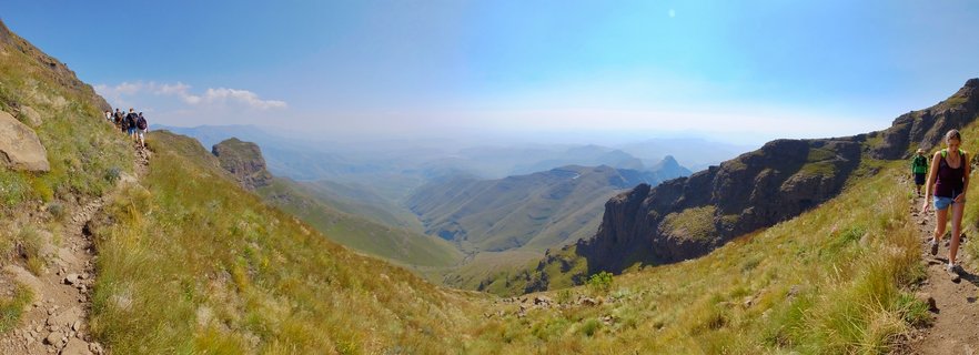 Panoramic view of hikers on a mountain trail in the Drakensberg with grassy slopes, rocky cliffs, and a deep valley stretching into the distance under blue sky