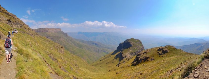 Panoramic view of hikers walking on a mountain trail in the Drakensberg range with green slopes, rocky peaks, and distant mountain ranges under a blue sky with scattered clouds