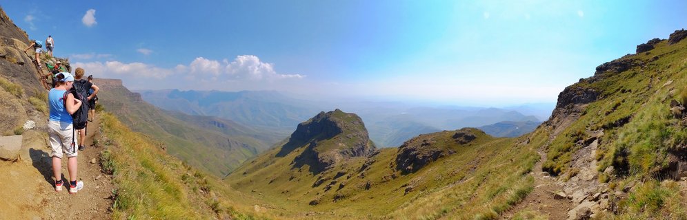 Group of hikers walking on a narrow dirt trail along a mountainside with expansive valley views and distant mountain peaks in the Drakensberg range