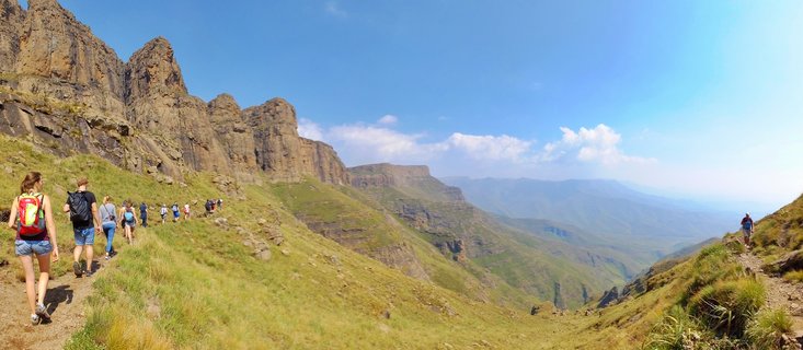 Group of hikers with backpacks walking on a mountain trail through grassy slopes with rocky cliffs and distant mountain ranges visible under blue sky