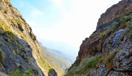 Steep rocky cliff faces on both sides frame a view through a mountain pass to a hazy valley and distant mountain ridges in the Drakensberg