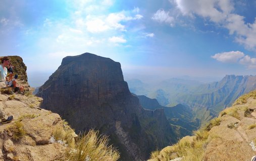 Mountain landscape showing rocky cliff edge with hikers on the left, dramatic peak in center, and layered mountain ranges extending into distant valleys under blue sky with white clouds