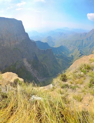 Mountain landscape showing steep rock cliffs and deep valleys with golden grasses in foreground and blue sky above in Northern Drakensberg South Africa