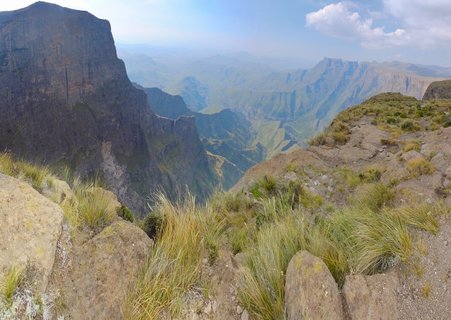 Mountain landscape showing steep rocky cliffs on the left side with a deep valley extending into the distance, foreground features rocky terrain with yellow-green tussock grasses under a partly cloudy sky
