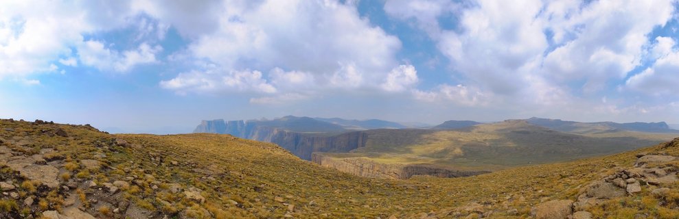 Wide mountain landscape showing rocky grassland foreground leading to dramatic vertical cliff faces and distant mountain ranges under blue sky with white clouds
