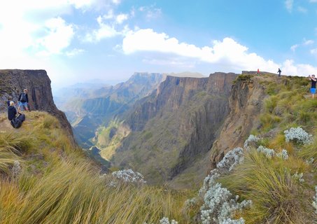 Wide mountain landscape showing hikers standing on grassy cliff edges overlooking a deep canyon with steep rock faces and distant mountain ranges under blue sky with white clouds