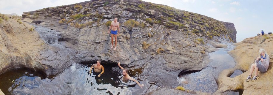 People swimming and jumping into natural rock pools in a mountainous landscape with rocky cliffs and sparse vegetation