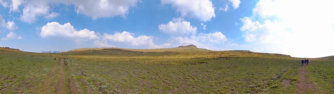 Wide panoramic landscape of Northern Drakensberg highland grasslands in South Africa showing rolling green and golden hills under blue sky with white cumulus clouds, dirt tracks winding through terrain, small group of hikers visible on right side, distant structures on left horizon