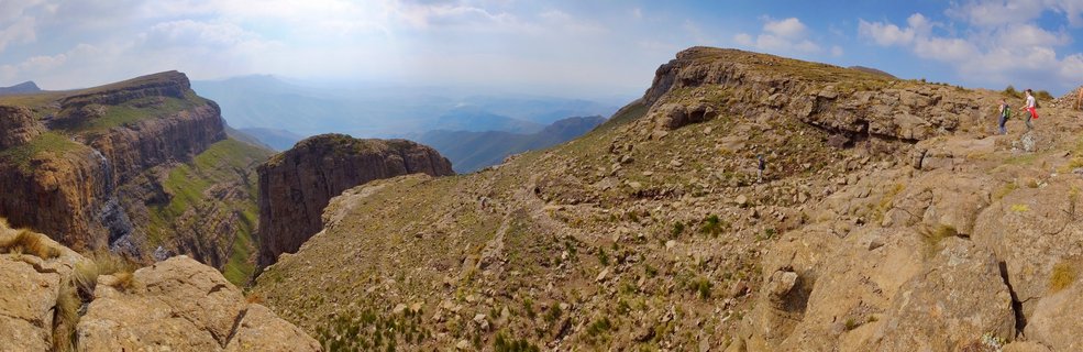 Panoramic view of hikers on rocky mountain terrain in Northern Drakensberg, South Africa, with steep cliffs, green valleys, and distant mountain ranges under blue sky with clouds