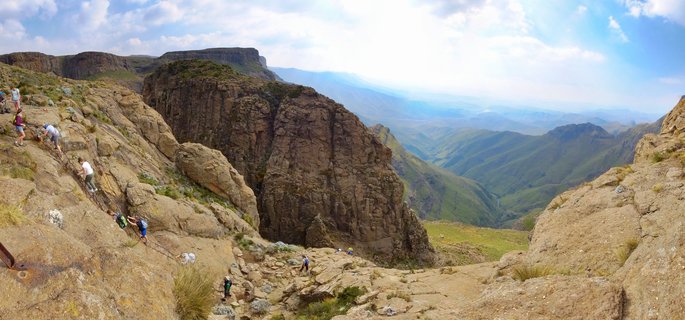 Multiple hikers with backpacks climb a steep rocky mountain pass using safety chains, with dramatic valley and distant mountain range views in the Northern Drakensberg, South Africa