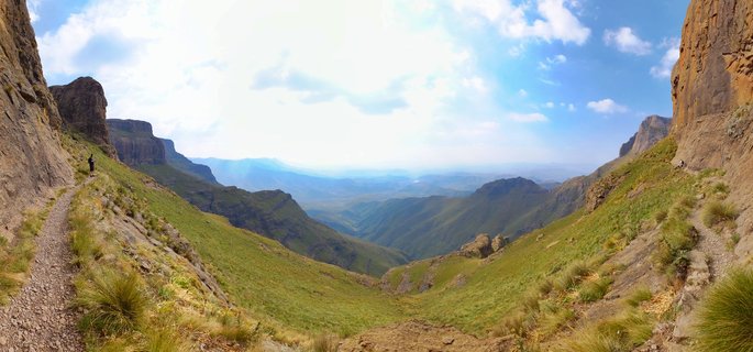 Panoramic view of a mountain valley in Drakensberg with steep rocky cliffs on both sides, green grassy slopes, a hiking trail with a small hiker visible on the left, and distant mountain ranges under blue sky with white clouds