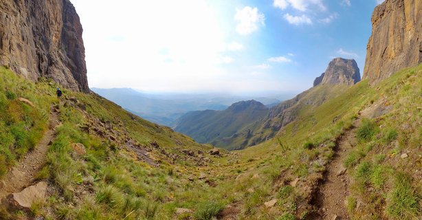 Mountain landscape showing a hiker on a dirt trail between tall rocky cliffs with green grassy slopes and distant mountain ranges under blue sky