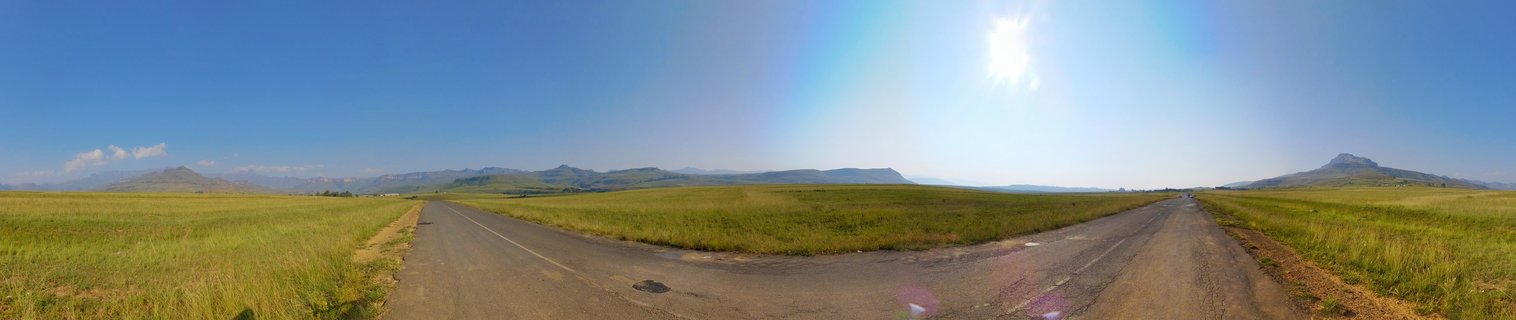 A Y-shaped road intersection through grassy plains with mountains in the distance under a bright blue sky in South Africa's Drakensberg region
