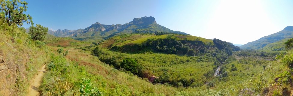 Panoramic landscape showing green valley with hiking trail on the left, stream running through center, and rocky mountain peaks in the background under blue sky in Northern Drakensberg, South Africa