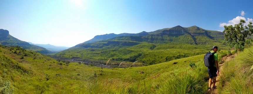 A person wearing a green shirt and backpack hiking on a dirt trail through green mountainous landscape with large mountains in the background under blue sky