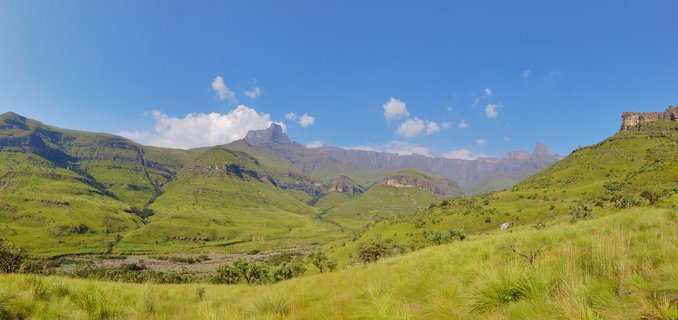 Panoramic view of green rolling hills leading to dramatic cliff-faced mountains under blue sky with scattered white clouds in Northern Drakensberg, South Africa