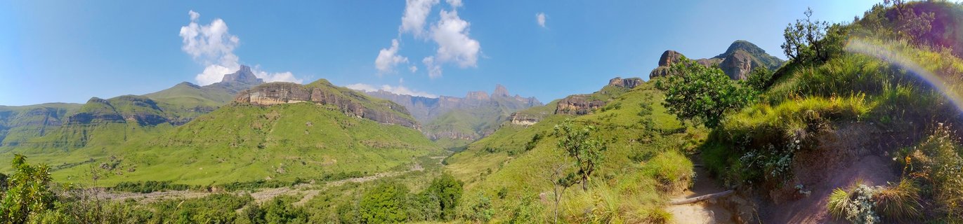 Panoramic landscape showing green mountain valleys with rocky Drakensberg peaks in the background, blue sky with white clouds, and a hiking trail on the right side
