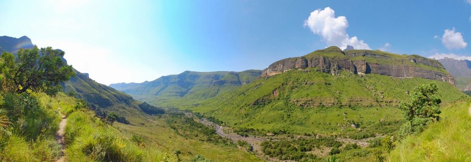 Panoramic landscape showing green Drakensberg mountains with rocky cliffs, a hiking trail on the left, valley with dry riverbed, blue sky with white clouds, South Africa