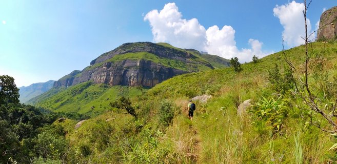 A hiker wearing a backpack walks along a narrow dirt trail through green mountain grassland, with large rocky cliff faces and mountains in the background under a blue sky with white clouds.