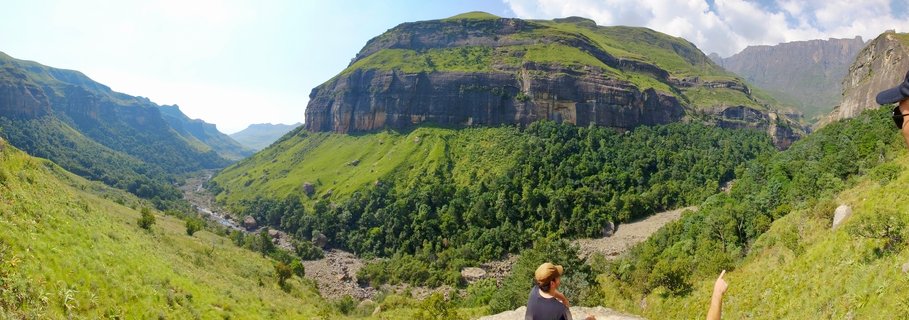Person sitting on rock ledge overlooking green mountain valley with steep cliff faces and river in Northern Drakensberg, South Africa