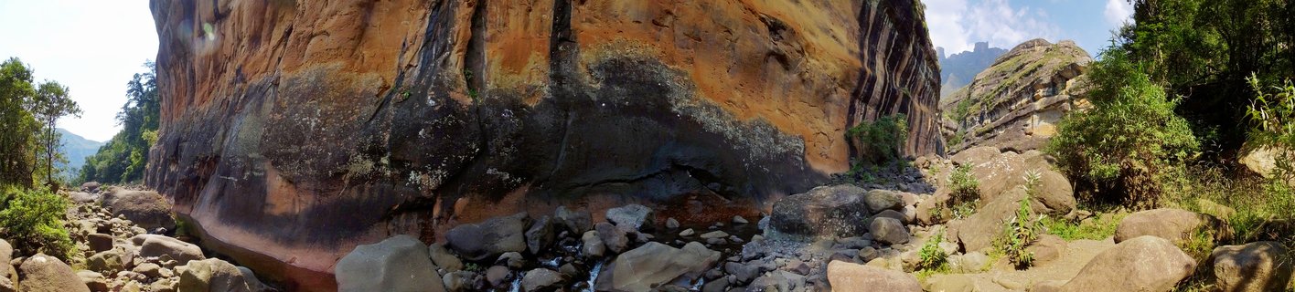 Large orange-brown sandstone cliff face with dark water stains towering over a rocky stream bed with boulders and green vegetation in Northern Drakensberg mountains