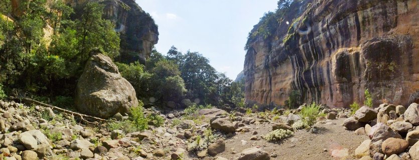Panoramic view of a rocky gorge in the Northern Drakensberg mountains showing tall sandstone cliff walls with dark vertical water stains, a large boulder in the foreground, scattered stones across a dry riverbed, green trees and vegetation on the left side and cliff tops, and clear blue sky above