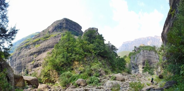 Mountain landscape showing layered sandstone cliffs and rock formations with green trees and vegetation in a valley with boulders and a hiker in the Northern Drakensberg, South Africa