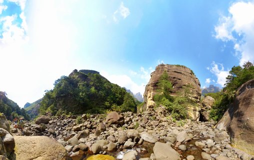 Wide landscape showing large sandstone rock formations and cliffs rising above a rocky riverbed with boulders, green vegetation on hillsides, blue sky with white clouds, and hikers on a trail at left