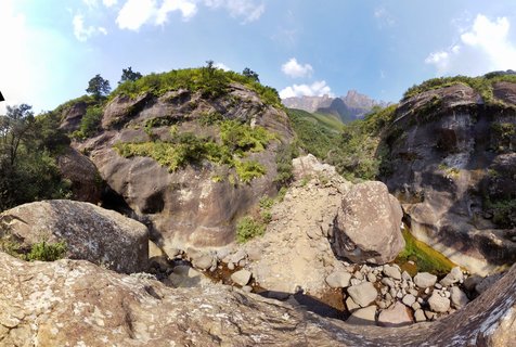Large weathered boulders and rock formations in a mountain gorge with green vegetation, smaller stones in a dry stream bed, and mountain peaks visible in the distance under blue sky with clouds.