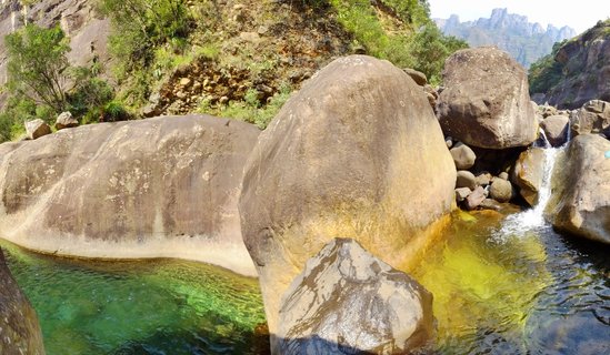 Large smooth granite boulders sit in a clear mountain stream with greenish-yellow water, a small waterfall cascades between rocks, with vegetated hillsides and distant mountains in the background