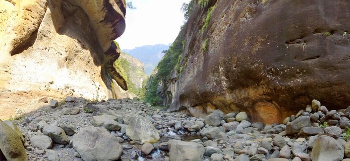 Rock canyon with large sandstone cliff walls on both sides, boulder-covered ground with small stream, green vegetation on rock faces, mountains visible in distance