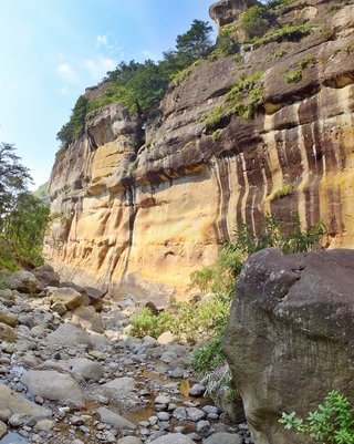 Tall sandstone cliff with dark vertical water stains rising above a rocky riverbed with boulders and green vegetation in Northern Drakensberg