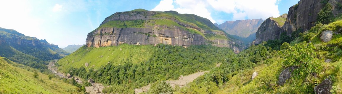 Panoramic view of green mountain valley in Drakensberg with rocky cliffs, hiking trail, and a person in green shirt walking on the right side under blue sky with white clouds