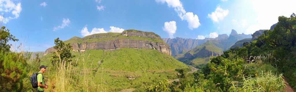 A hiker wearing a green shirt and backpack stands on a mountain trail looking at the Drakensberg mountain range with green slopes and layered rock cliffs under a blue sky with white clouds