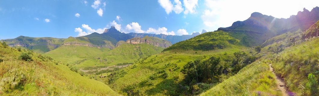 Wide mountain landscape showing green rolling hills in the foreground with dramatic rocky Drakensberg mountain peaks in the background under a blue sky with white clouds, hiking trail visible on the right side