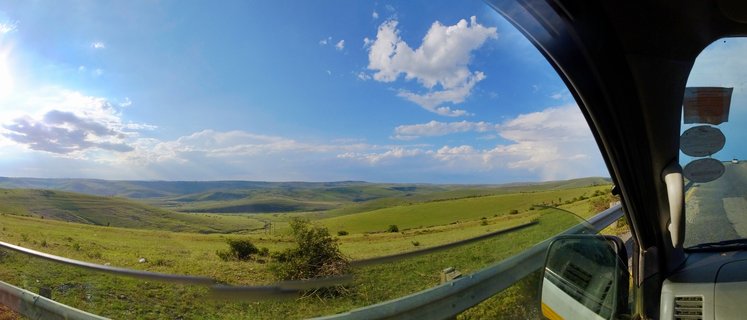 View of rolling green hills and valley landscape in Coffee Bay, South Africa, photographed from inside a vehicle with guardrail visible in foreground under blue sky with scattered clouds