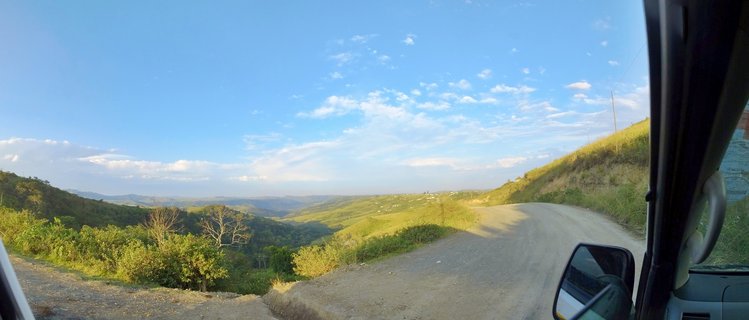 View from inside a vehicle showing a winding dirt road through green rolling hills under a blue sky with white clouds in Coffee Bay, South Africa