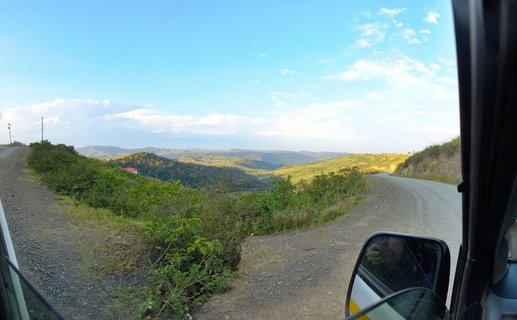 A winding gravel road curves through green rolling hills with blue sky and scattered clouds, viewed from inside a vehicle with side mirror visible in foreground