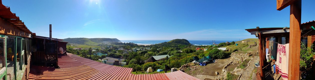 Wide panoramic photograph showing a coastal village with red metal rooftops in the foreground, green rolling hills with scattered houses, and blue ocean visible in the distance under a clear sky