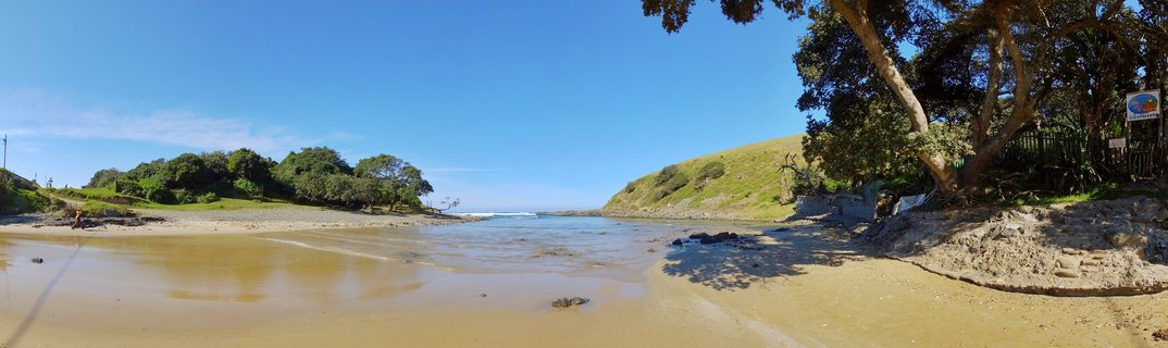 River estuary flowing across sandy beach to ocean with green hills and trees under blue sky at Coffee Bay, South Africa