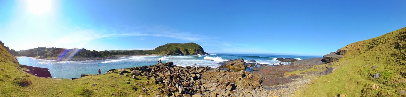 Panoramic coastal landscape showing rocky shoreline with waves, green grassy hills, and clear blue sky at Coffee Bay, South Africa
